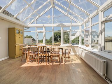 A conservatory with a table and wooden chairs and a yellow storage cabinet at Auverne in Poundstock near Widemouth Bay