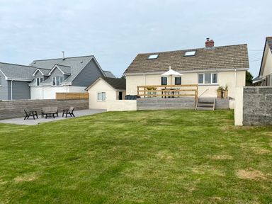 A backyard with a large lawn a raised wooden deck with table chairs and umbrella and a paved patio with outdoor seating at Auverne Poundstock near Widemouth Bay