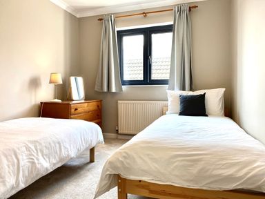 A bedroom with two single beds a wooden dresser with a lamp and mirror and a window with curtains at Auverne Poundstock near Widemouth Bay