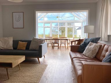 A living room with two sofas and a wooden table leading to a dining area with wooden chairs in a glass conservatory at Auverne in Poundstock near Widemouth Bay