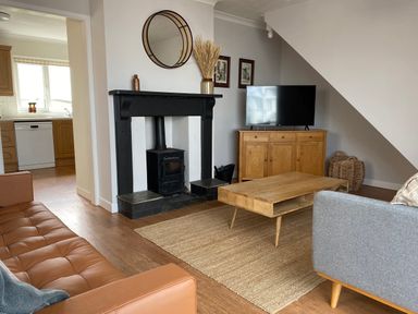 A living room with a wood stove fireplace a brown leather sofa a wooden coffee table a gray armchair and a wooden cabinet with a TV at Auverne Poundstock near Widemouth Bay