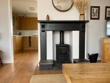 A living room with a black wood burning stove and wooden furniture leading to a kitchen with wooden cabinets at Auverne in Poundstock near Widemouth Bay