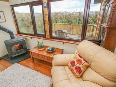 A living room with a leather sofa a wood burning stove a wooden table and large windows overlooking a wooden deck with chairs at Sarngwm in Narberth