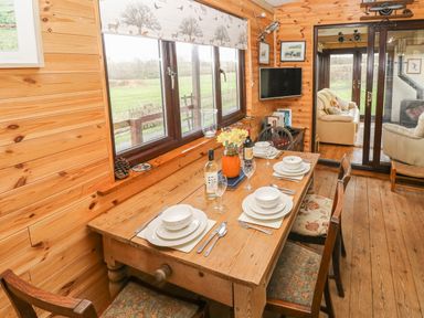 A dining area with a wooden table set for four next to windows with a countryside view and a living room visible through glass doors at Sarngwm in Narberth