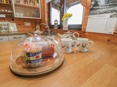A wooden kitchen counter with two mugs a teapot a glass cake cover with cakes under it and a calendar on the wall at Sarngwm in Narberth