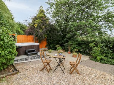 An outdoor seating area with a small wooden table and three wooden chairs on gravel near a hot tub and surrounded by greenery at Fisherman's Cottage in Llandysul