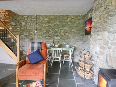 A room with a stone wall a staircase an orange armchair a small dining table with chairs and a wood stove with a basket of firewood at Fisherman's Cottage in Llandysul