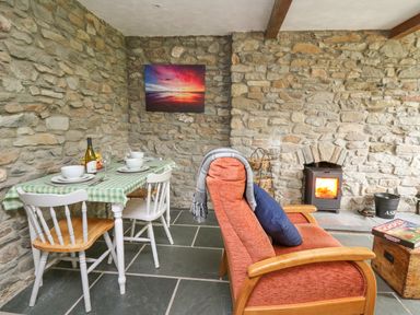 A dining table with chairs and a wine bottle next to an armchair and a stove in a stone wall room at Fisherman's Cottage in Llandysul
