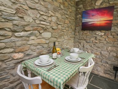 A dining table with a checkered tablecloth set with bowls plates utensils a bottle of wine and a fruit basket in a room with stone walls and a colorful sunset picture at Fisherman's Cottage in Llandysul