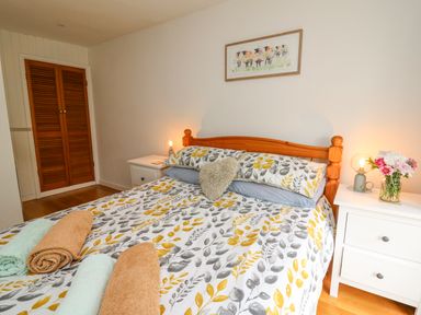 A bedroom with a wooden bed, floral bedding, two bedside tables with lamps and flowers, and a closed wooden closet door at Fisherman's Cottage in Llandysul