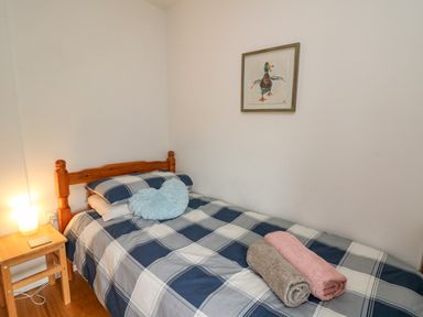 A bedroom with a single bed with checkered bedding two rolled towels a fluffy heart-shaped pillow a wooden bedside table with a lamp and a duck painting on the wall at Fisherman's Cottage in Llandysul