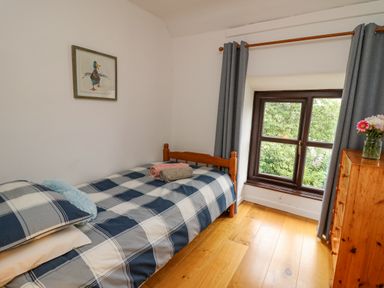 A bedroom with a single bed with blue check bedding a window with grey curtains and a wooden chest of drawers with flowers at Fisherman's Cottage in Llandysul
