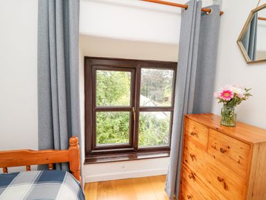 A wooden bed with a blue and white striped blanket near a window with grey curtains and a wooden dresser with flowers in a glass jar at Fisherman's Cottage in Llandysul