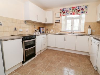 A kitchen with white cabinets, a black oven, a sink below a window with floral curtain, and beige tiled floor at The Vale in Ashwater