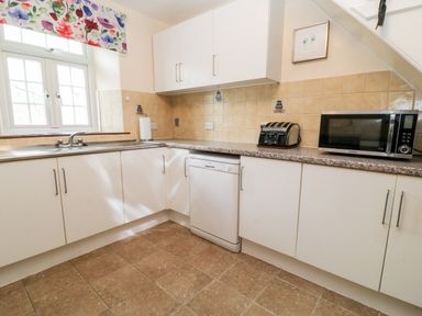 A kitchen with white cabinets a dishwasher a toaster a microwave and a window with floral curtains at The Vale in Ashwater