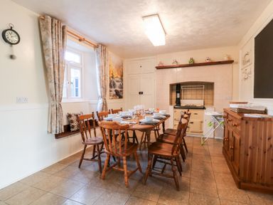 A dining room with a wooden table and chairs set with dishes next to a window with curtains and a tiled stove at The Vale in Ashwater