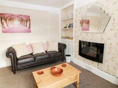 A living room with a brown leather sofa a wooden coffee table with bowls a wall-mounted electric fireplace and a built-in shelving unit at The Vale in Ashwater