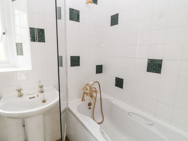 A bathroom with a white bathtub and sink with gold fixtures and tiled walls with green square accents at The Vale in Ashwater