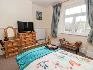 A bedroom with a wooden bed a chest of drawers with a television a small wooden dresser a window with curtains and a colorful bench at The Vale in Ashwater