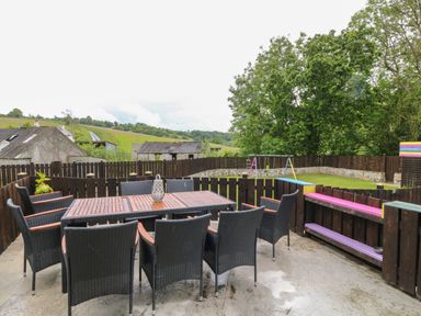 An outdoor patio with a table and eight chairs next to a fenced grassy area with a swing set at The Vale in Ashwater