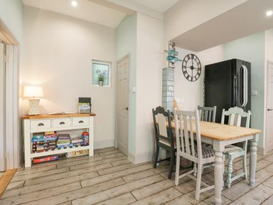 A dining area with a wooden table and mismatched chairs next to a black refrigerator and a side table with board games and a lamp at Saxon Lodge Cottage Felixstowe Suffolk