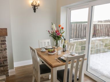 A dining area with a wooden table set for four with chairs near a large sliding glass door at Shipswheel Cottage at Broadings Farm in Whitby