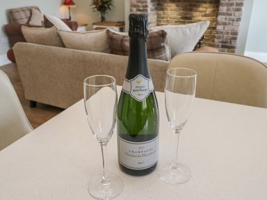 A bottle of champagne with two empty champagne flutes on a table in a living room with a sofa and a brick fireplace at Shipswheel Cottage at Broadings Farm Whitby
