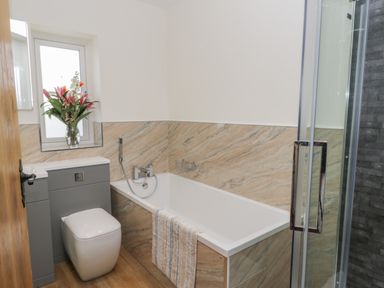 A bathroom with a bathtub a toilet a vase with flowers on the window sill and a glass shower door at Shipswheel Cottage at Broadings Farm in Whitby
