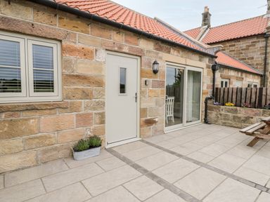 A stone cottage exterior with a beige door window and sliding glass door leading to a patio with a wooden picnic table and planter boxes at Shipswheel Cottage at Broadings Farm Whitby