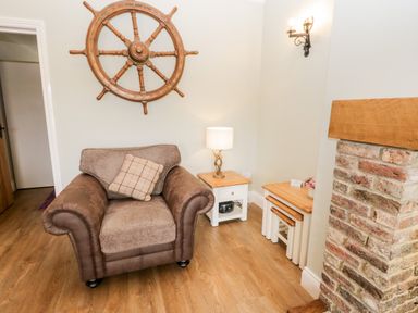 A brown armchair with a cushion beside a wooden side table with a lamp and nesting tables near an exposed brick fireplace at Shipswheel Cottage at Broadings Farm Whitby