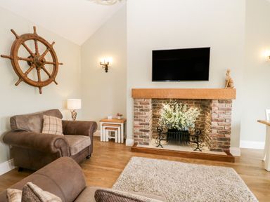 A living room with a fireplace TV armchairs and a wooden ship wheel on the wall at Shipswheel Cottage at Broadings Farm Whitby