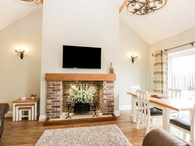A living room with a brick fireplace white flowers a wall mounted television and a dining table with chairs at Shipswheel Cottage at Broadings Farm in Whitby