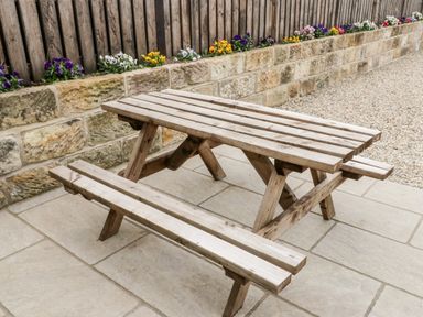 A wooden picnic table on a stone patio next to a stone wall with a flower bed and wooden fence at Shipswheel Cottage at Broadings Farm in Whitby