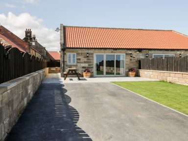 An exterior view of a stone building with a red tiled roof a paved area a wooden picnic table two flower pots a lawn and wooden fencing at Stag Cottage at Broadings Farm in Whitby