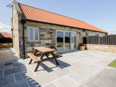 A stone building with a red tiled roof a wooden picnic table and flower pots on a paved patio at Stag Cottage at Broadings Farm Whitby