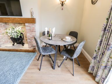 A dining area with a round table set with plates glasses and a bottle next to a brick fireplace and floral curtains at Stag Cottage at Broadings Farm in Whitby