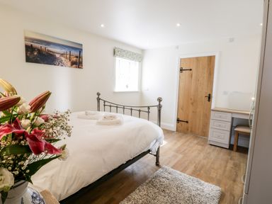 A bedroom with a metal bed frame white bedding a wooden door a small desk with a chair flowers on a bedside table and a window at Stag Cottage at Broadings Farm Whitby
