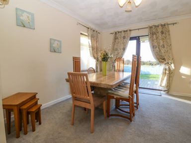 A dining room with a wooden table and six chairs with cushions a vase with flowers on the table curtains and glass doors leading outside at Rhiangwyn in Llanrhyddlad near Cemaes Bay