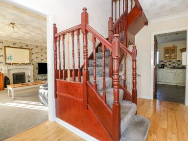 A hallway with wooden stair railing and carpeted stairs with a living room visible through an open door and a kitchen through another door at Rhiangwyn in Llanrhyddlad near Cemaes Bay