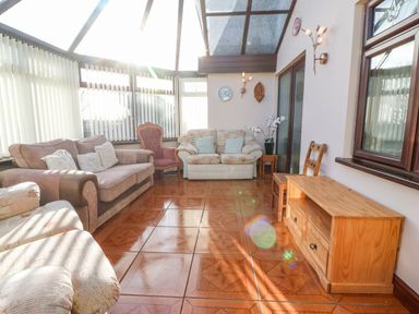 A sunlit conservatory with sofas a wooden chair a wooden cabinet and tiled floor at Rhiangwyn in Llanrhyddlad near Cemaes Bay