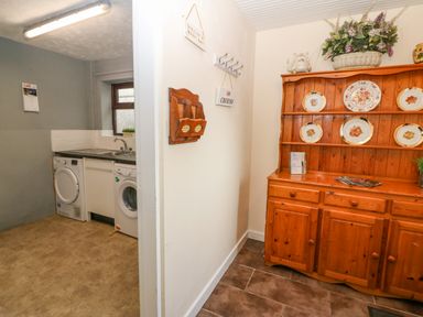 A hallway with a wooden cabinet holding plates and decorative items next to a utility room with a washing machine sink and dryer at Rhiangwyn in Llanrhyddlad near Cemaes Bay