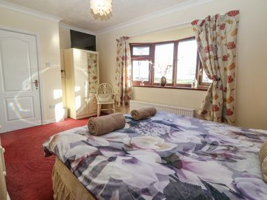 A bedroom with a bed with floral bedding two rolled towels on it a wardrobe a chair and a window with floral curtains at Rhiangwyn in Llanrhyddlad near Cemaes Bay