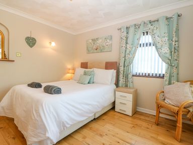 A bedroom with a bed cushions bedside table window with floral curtains and a wicker chair at Rhiangwyn in Llanrhyddlad near Cemaes Bay
