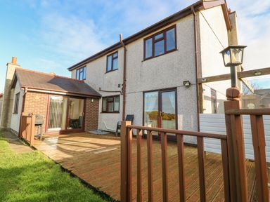 A backyard with wooden deck and lawn next to a two-story house with sliding glass doors and windows at Rhiangwyn in Llanrhyddlad near Cemaes Bay
