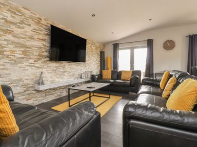 A living room with black leather sofas and yellow cushions with a mounted TV on a stone wall at Box Tree Escapes in Kirkby Lonsdale