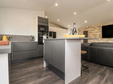 A kitchen with a dark grey island and cabinets a yellow kettle and bottle with glasses on the counter and a black sofa in a living area at Box Tree Escapes Kirkby Lonsdale