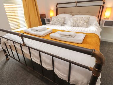 A bedroom with a metal bed frame white linens mustard yellow blanket towels on the bed two nightstands with lamps and mustard yellow curtains at Box Tree Escapes in Kirkby Lonsdale