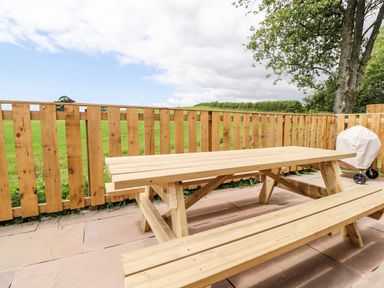 A wooden picnic table and benches on a stone patio with a wooden fence and green field background at Box Tree Escapes in Kirkby Lonsdale