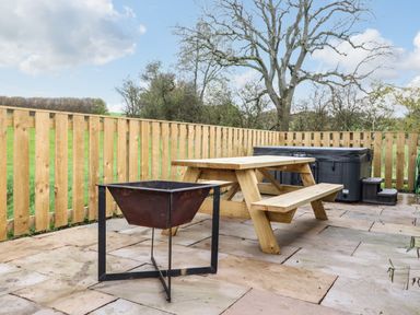 An outdoor patio with a wooden picnic table a fire pit and a hot tub surrounded by a wooden fence at Box Tree Escapes in Kirkby Lonsdale