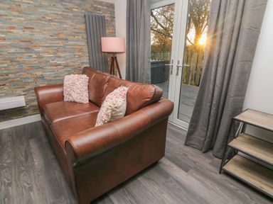 A living room with a brown leather sofa and two textured pillows next to glass doors with curtains and a small shelving unit at Box Tree Escapes Wennington near Kirkby Lonsdale
